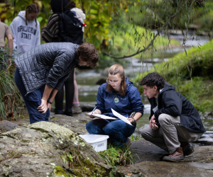 Three people examine papers and a plastic container while sitting on rocks beside a stream; others stand in the background near trees and water.